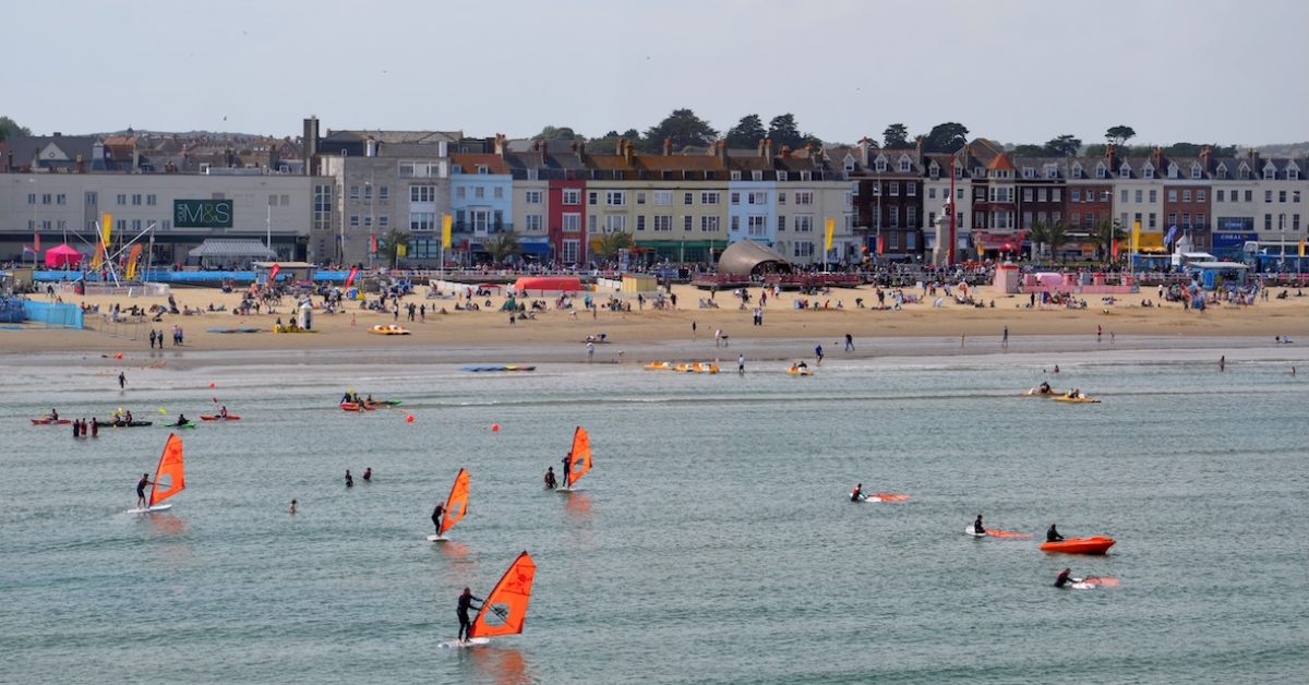 "Weymouth, UK - August 4, 2012: A view from the water to Weymouth seafront with people using windsurf boards and small boats. Many people on the sandy beach and the town behind."